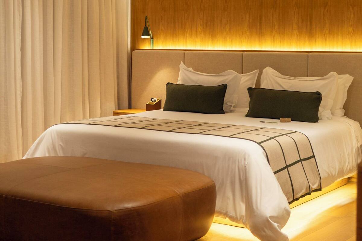 Un dormitorio de hotel moderno con cabecera de madera, una cama grande con ropa blanca, dos almohadas y una mesita de noche.