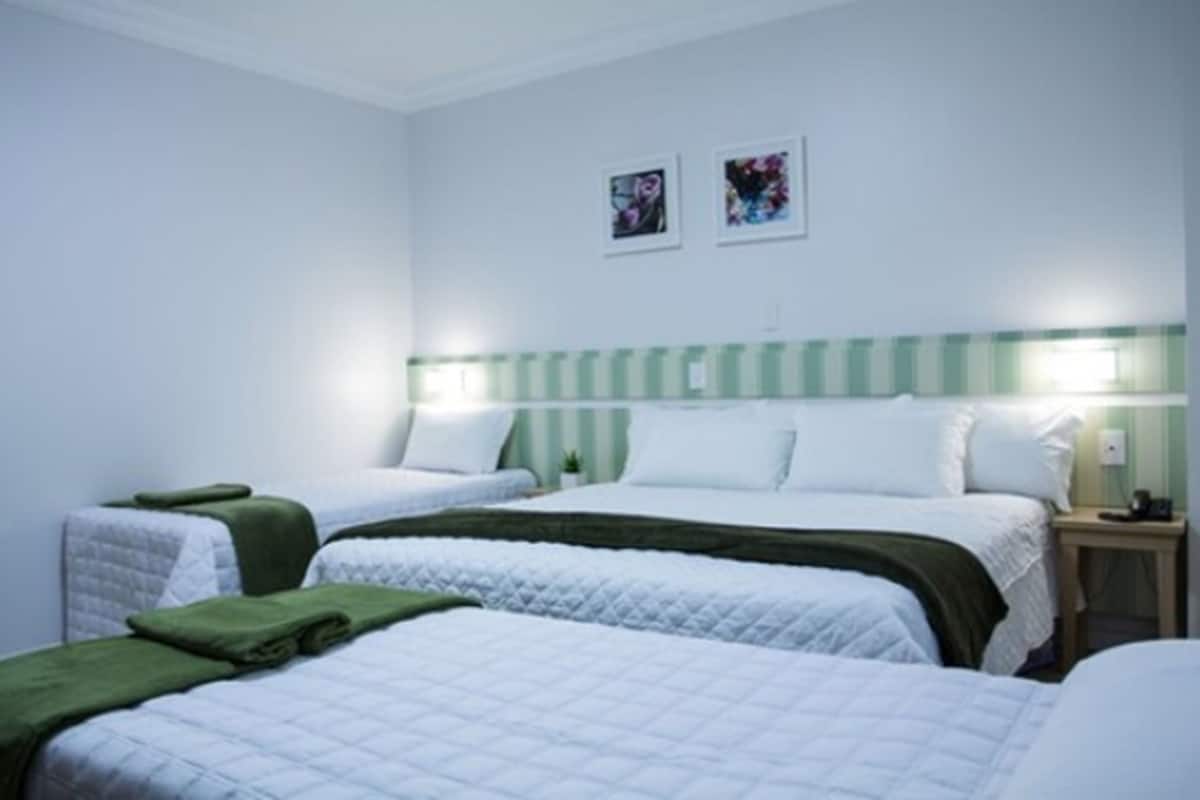 Habitación de hotel con dos camas, cada una con una manta verde y ropa de cama blanca. Hay dos mesitas de noche con lámparas. Las paredes están pintadas de blanco y hay dos cuadros enmarcados.