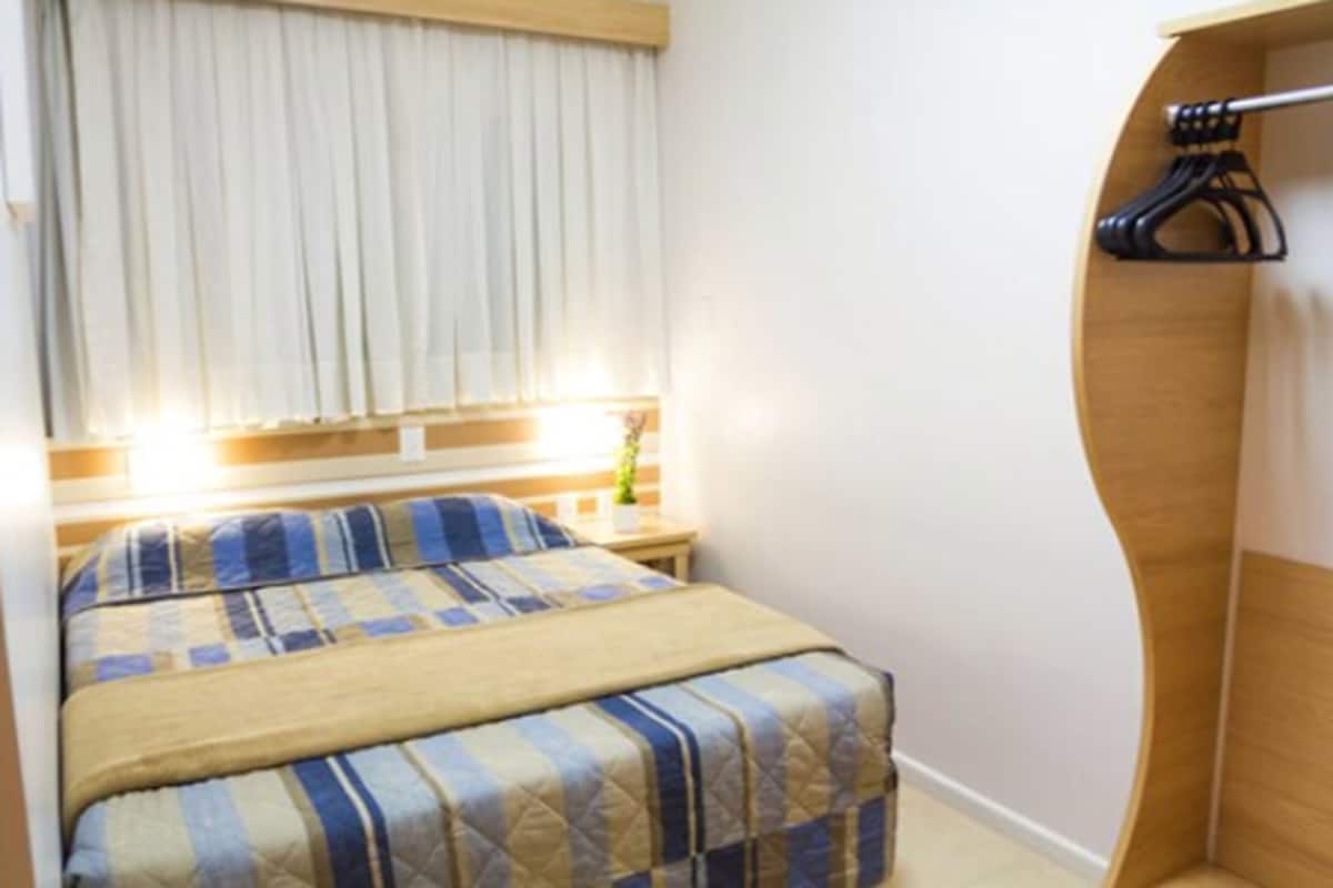 Una habitación de hotel con cama, mesita de noche, ventana con cortinas y un armario de madera.