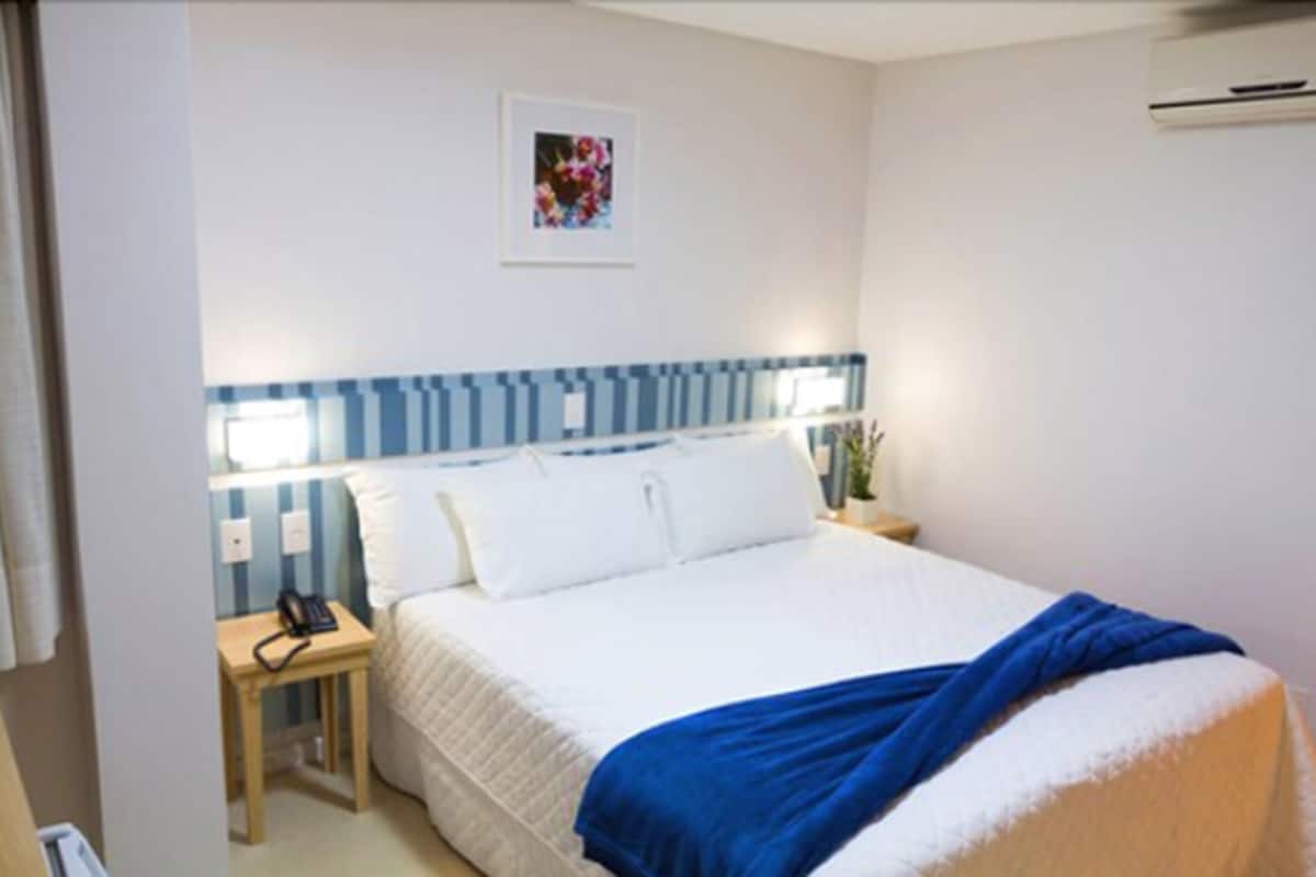 Una habitación de hotel con una cama, una mesita de noche con una lámpara, un aire acondicionado instalado en la pared y un cuadro enmarcado.