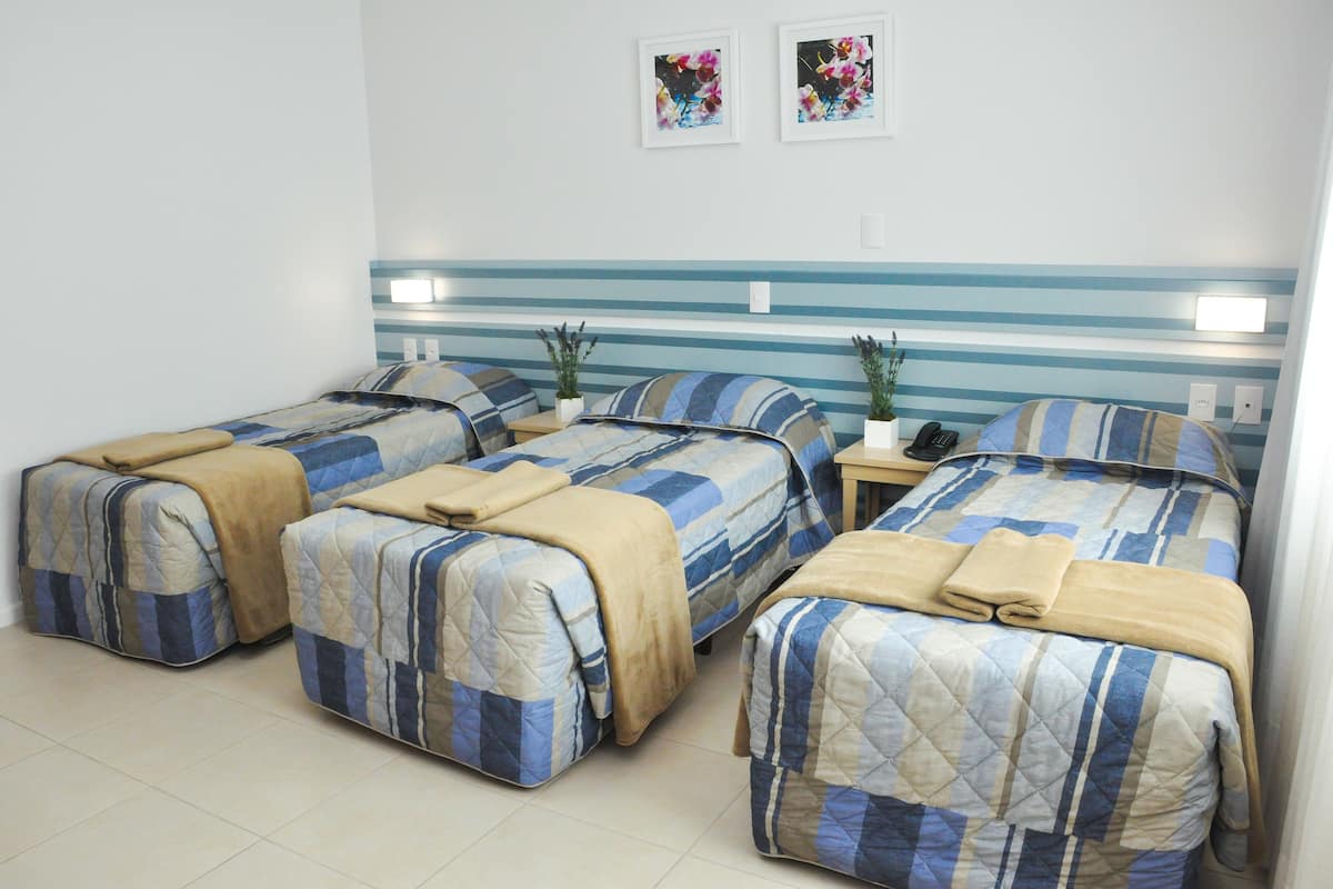 Habitación de hotel con tres camas, cada una con cabecero a rayas y una manta color beige. Hay dos lámparas de pared y dos cuadros con flores enmarcadas sobre las camas.