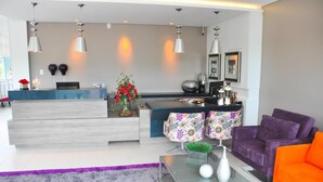 A modern living room with a reception desk, purple and orange furniture, and decorative items. - BR Suítes Hotel (Navegantes)