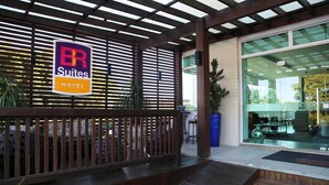 A hotel entrance with a sign for BR Suites Hotel, a covered outdoor area with wooden railings, and a glass door leading inside. - BR Suítes Hotel (Navegantes)