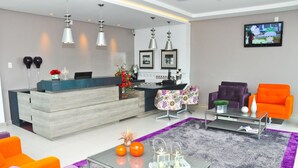 A modern living room with a reception desk, purple and orange sofas, a coffee table, and a TV mounted on the wall. - BR Suítes Hotel (Navegantes)