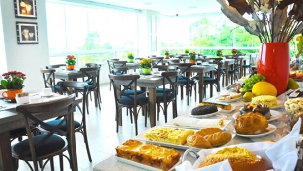 A dining area with tables set for a meal, featuring a variety of dishes and a vase with flowers. - BR Suítes Hotel (Navegantes)