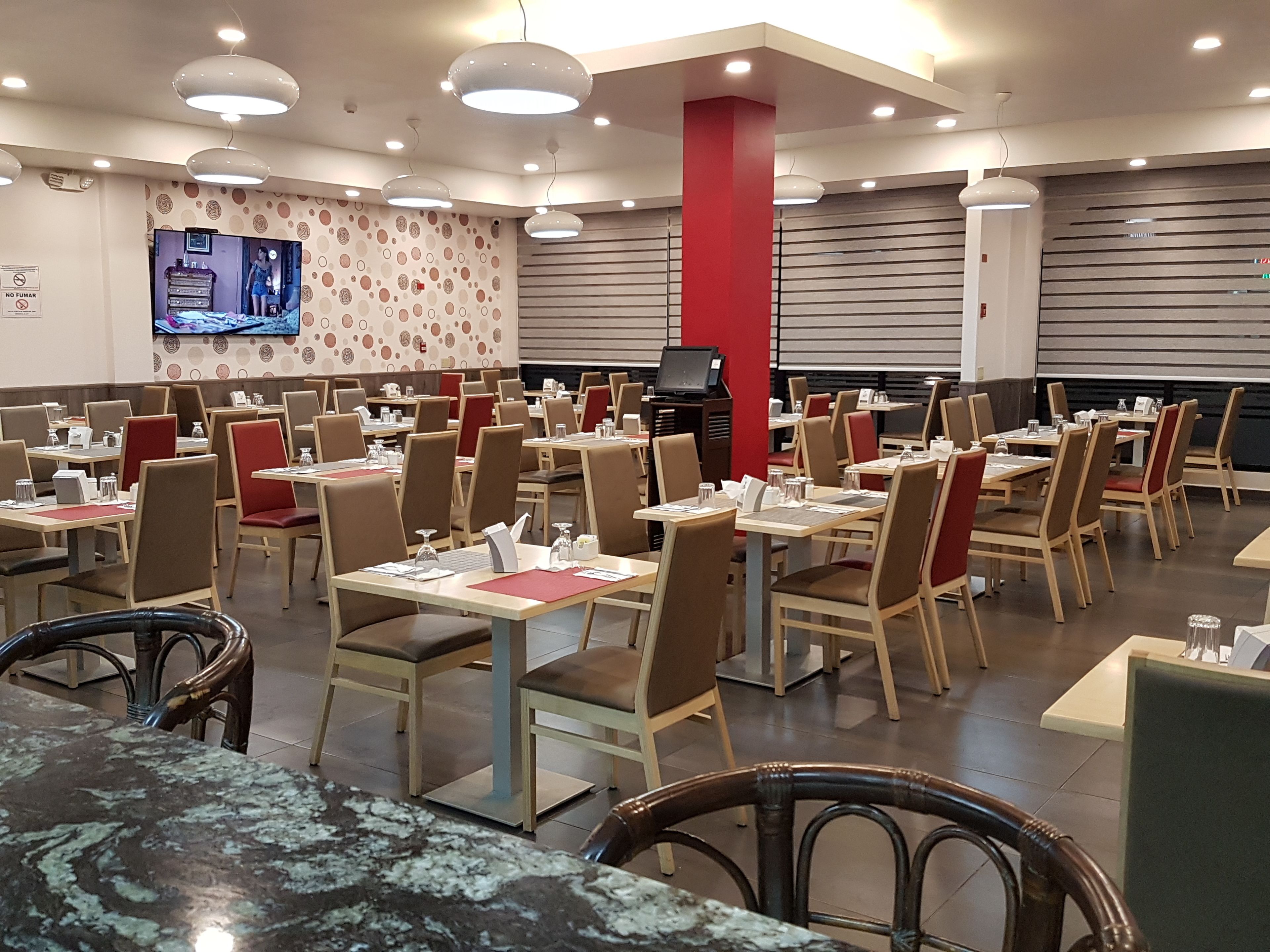 A restaurant with multiple tables and chairs, a television mounted on the wall, and a red pillar.
