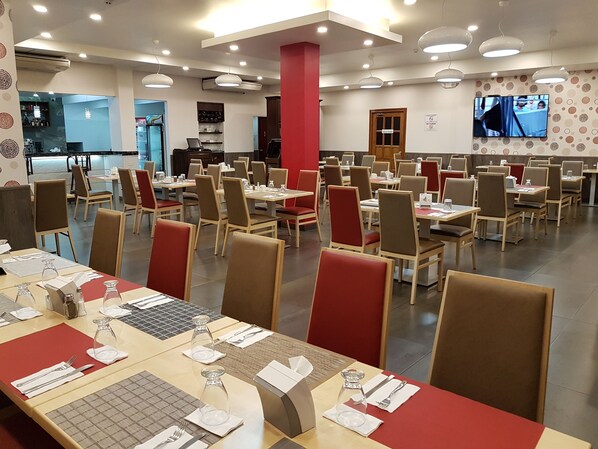A spacious restaurant with multiple tables and chairs, a television screen, and a counter area.