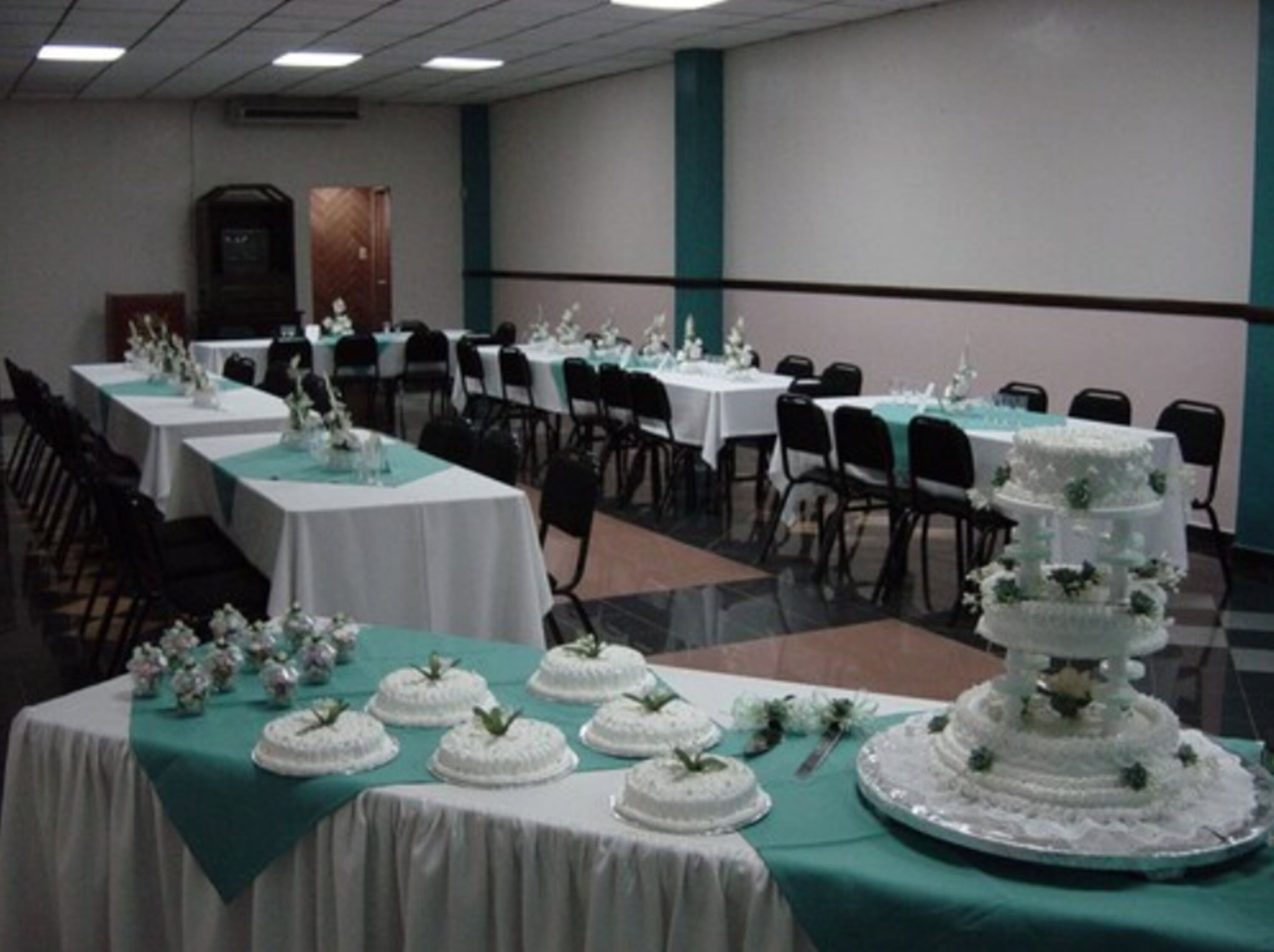 A banquet hall with multiple round tables covered with white tablecloths and teal table runners. There are several white tiered cakes on display, and the room has a modern design with a neutral color palette.