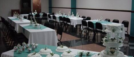 A banquet hall with multiple round tables covered with white tablecloths and teal table runners. There are several white tiered cakes on display, and the room has a modern design with a neutral color palette.