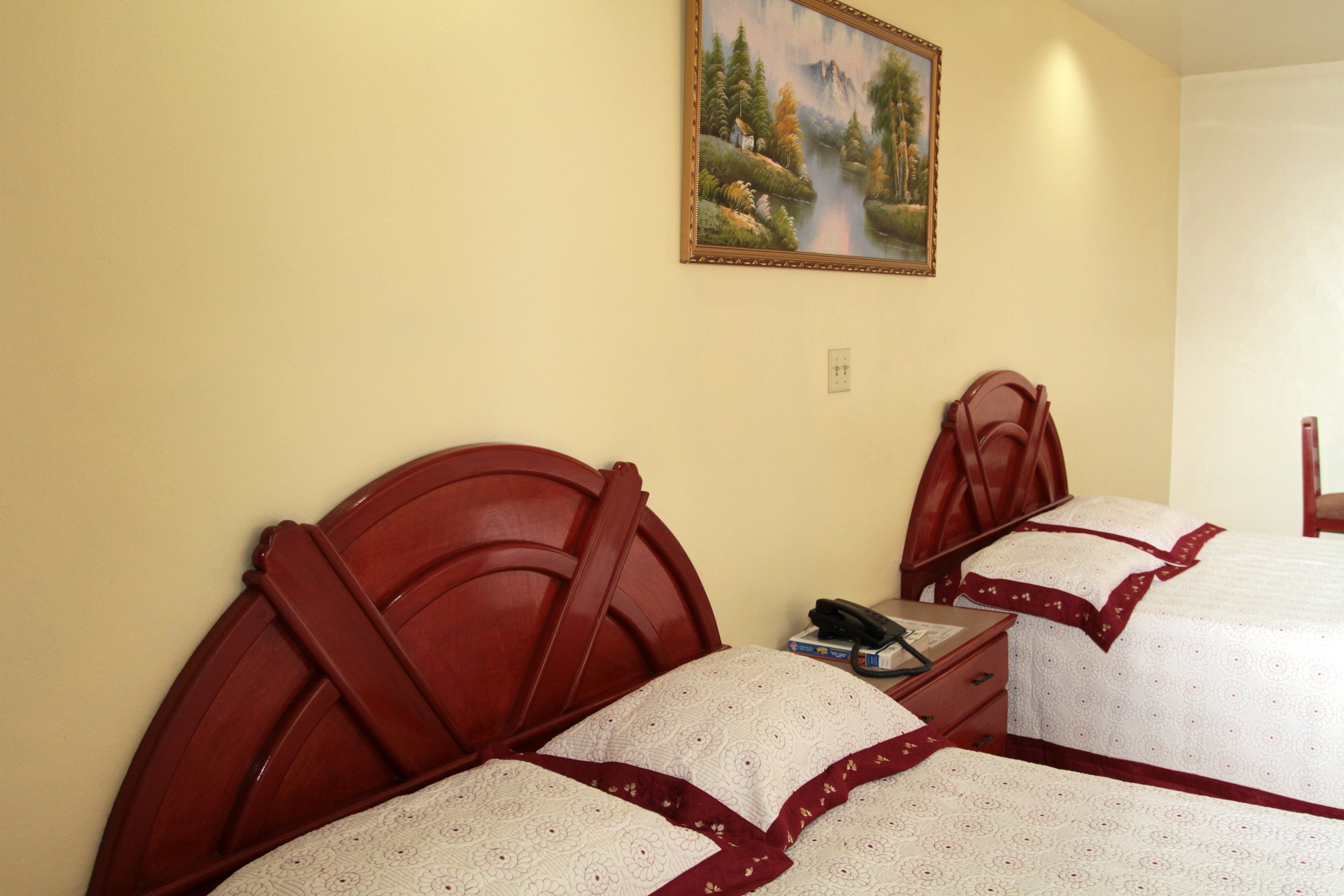 Une chambre d’hôtel avec deux lits, chacun doté d’une tête de lit en bois et de draps blancs agrémentés d’une bordure rouge. Un tableau représentant un paysage est accroché au mur, au-dessus des lits.