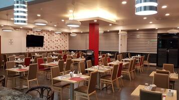 Un ristorante spazioso con numerosi tavoli e sedie, lampade moderne e un televisore a schermo piatto appeso al muro.