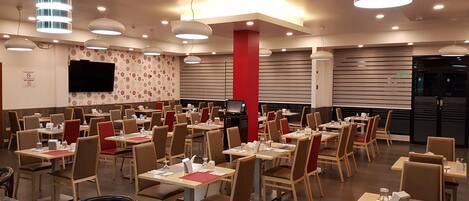 A spacious restaurant with multiple tables and chairs, modern lighting fixtures, and a flat-screen TV on the wall.