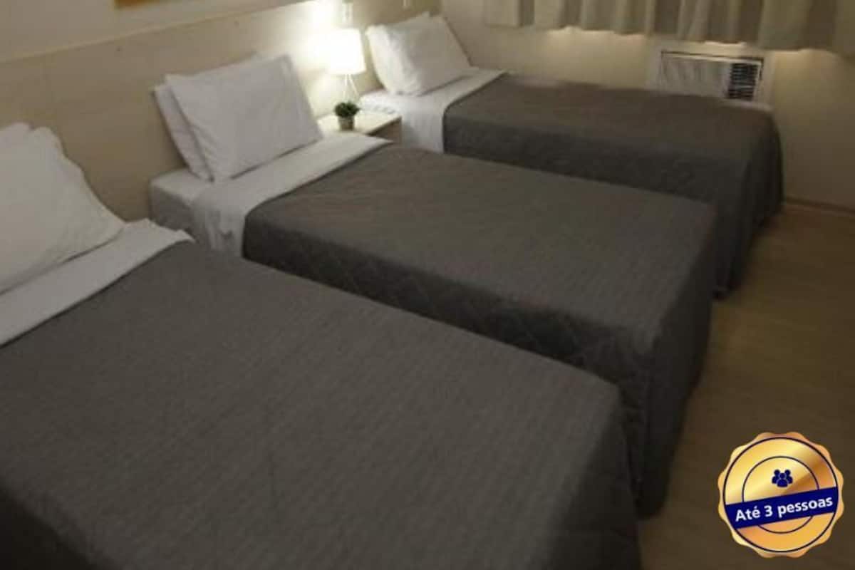 Una habitación de hotel con tres camas individuales, una mesita de noche con lámpara y un equipo de aire acondicionado.