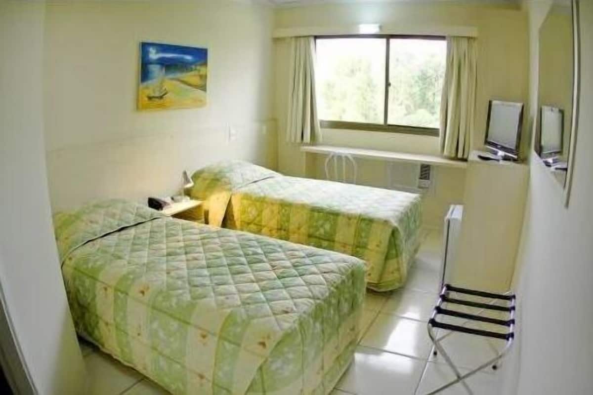 Una habitación pequeña y limpia con dos camas, una ventana con cortinas, un escritorio con computadora y un televisor en la pared.
