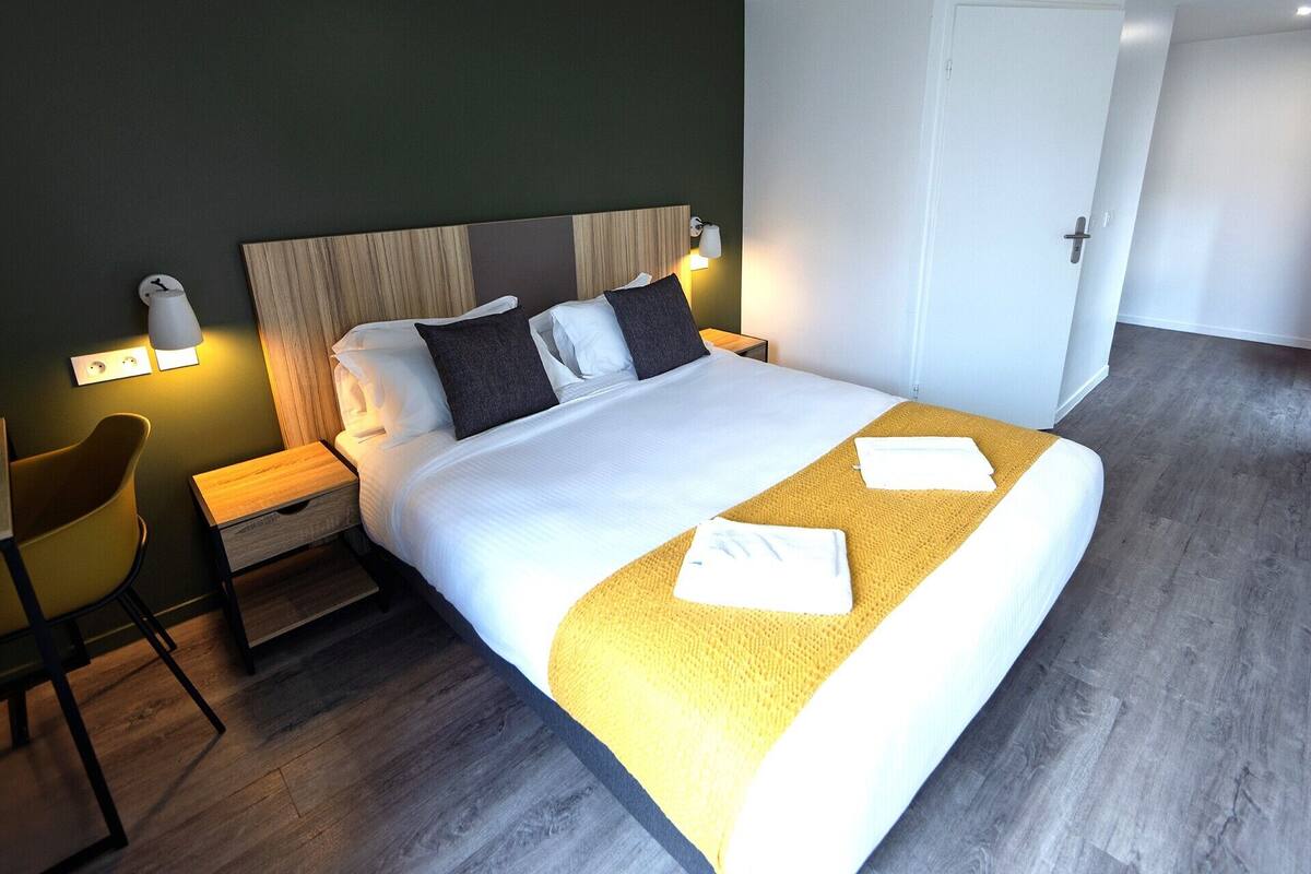 A neatly made bed with white linens and a yellow throw blanket, a wooden nightstand with a lamp, and a yellow chair.