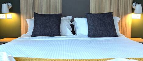 A hotel room with a large bed, two dark pillows, white pillows, a beige throw blanket, and two wall-mounted lamps.