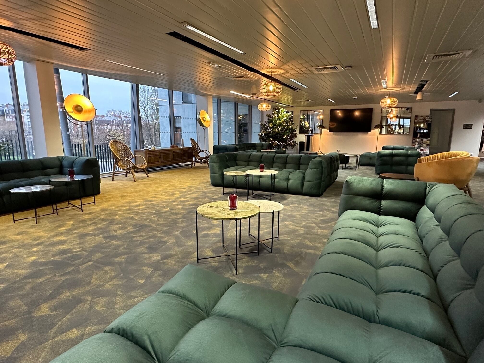 Et moderne loungeområde med grønne sofaer, runde borde og fletstole. Store vinduer giver udsigt over bybilledet.
