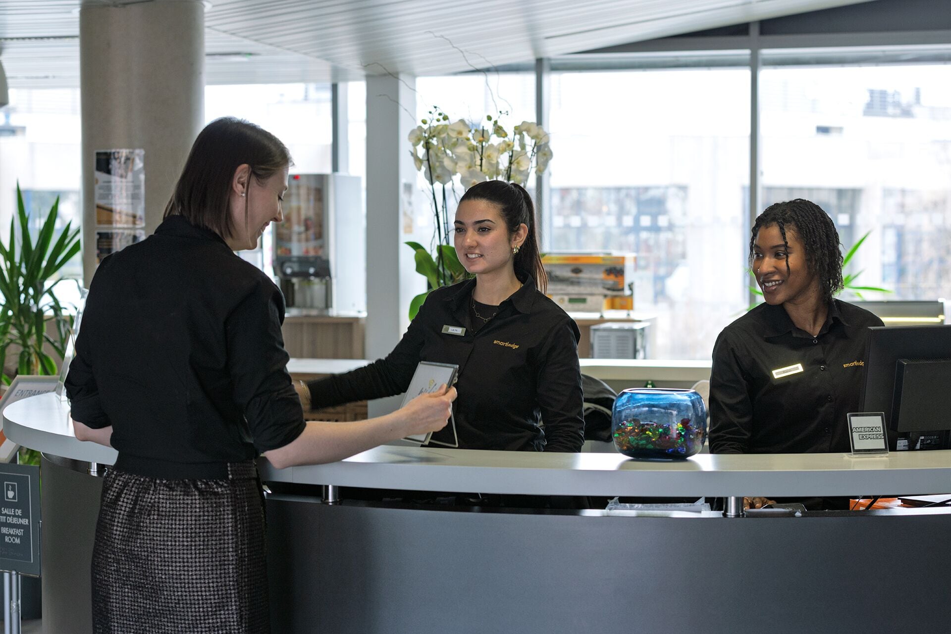A reception desk with three staff members in black uniforms. There is a sign indicating a breakfast room.