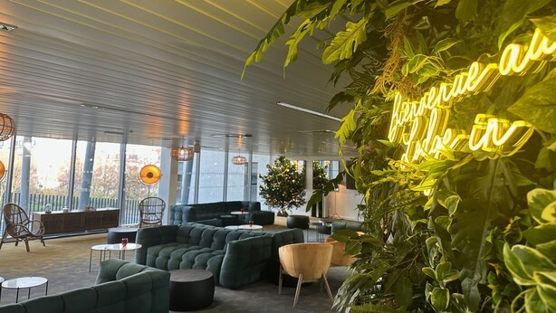 A modern lounge area with green sofas, a neon sign, and large windows.