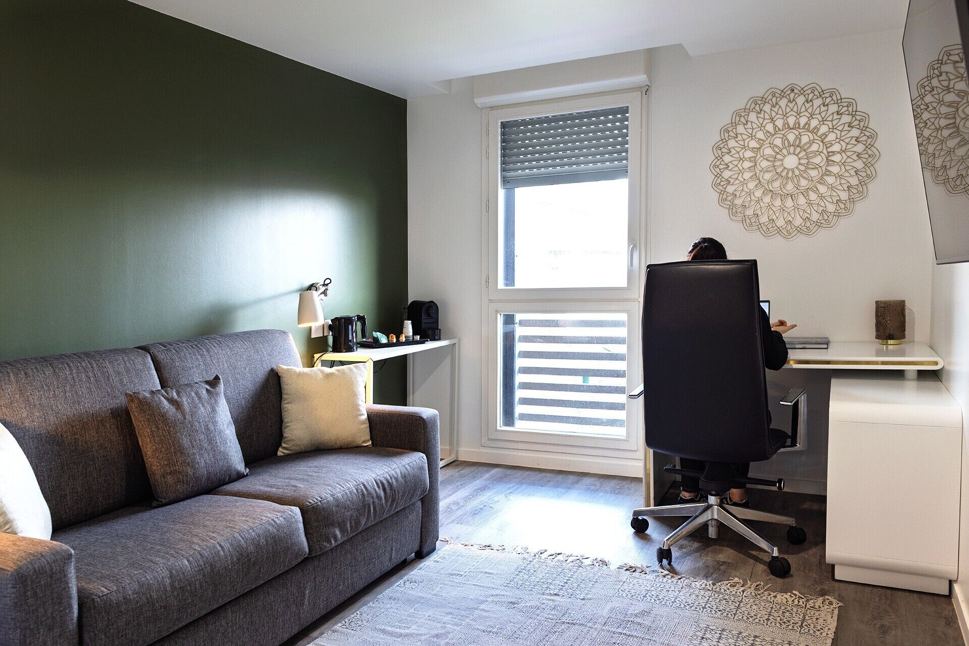 A modern living room with a grey sofa, a coffee table, a desk with a chair, and a large window with blinds.