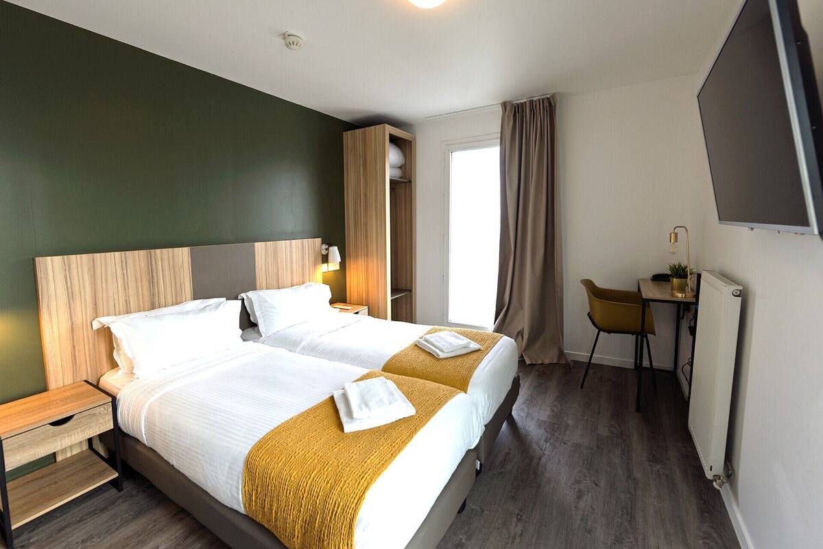 A hotel room with two beds, a desk, a chair, a TV, and a window with curtains.