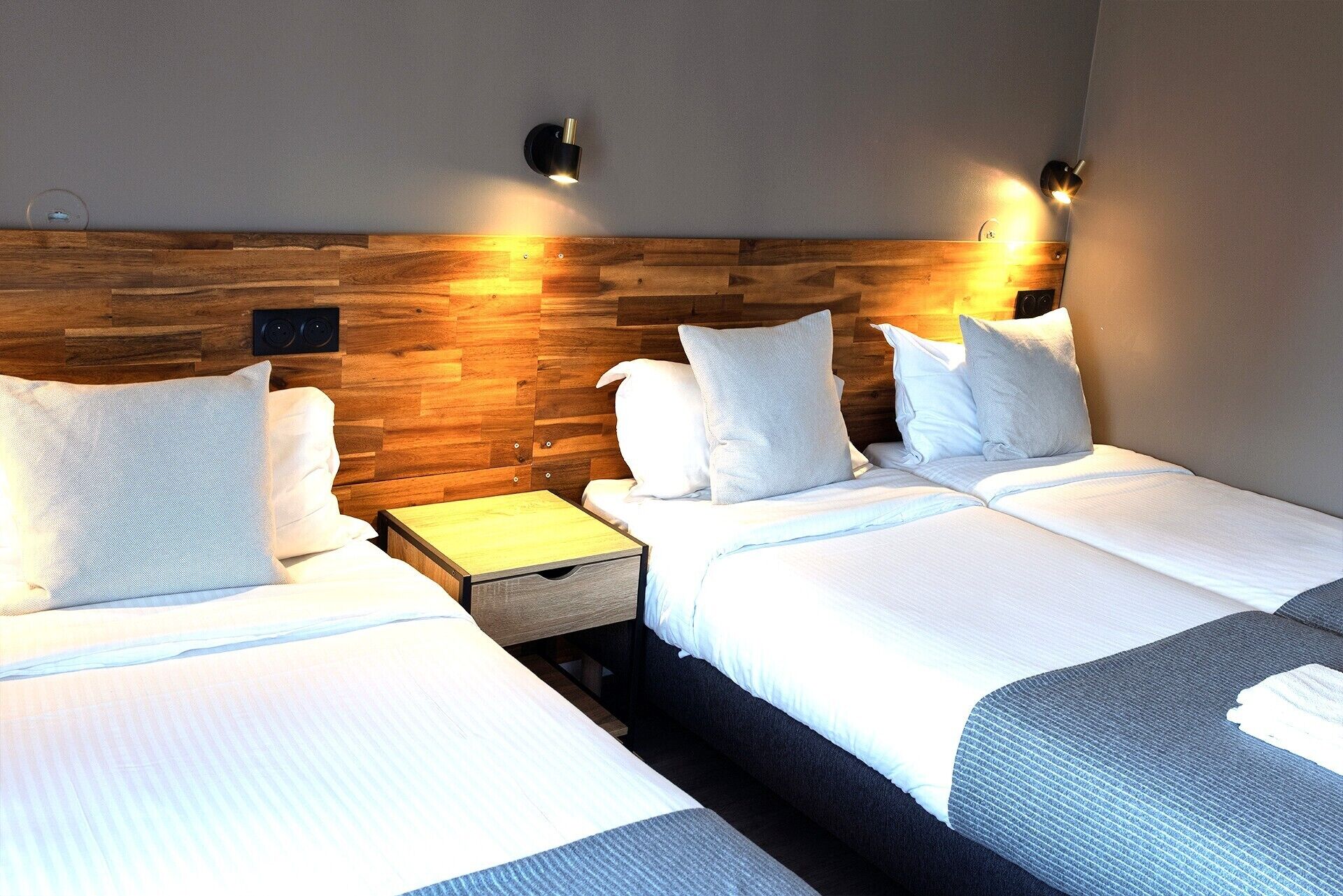 Habitación de hotel con dos camas, cabecero de madera, mesitas de noche e iluminación montada en la pared.