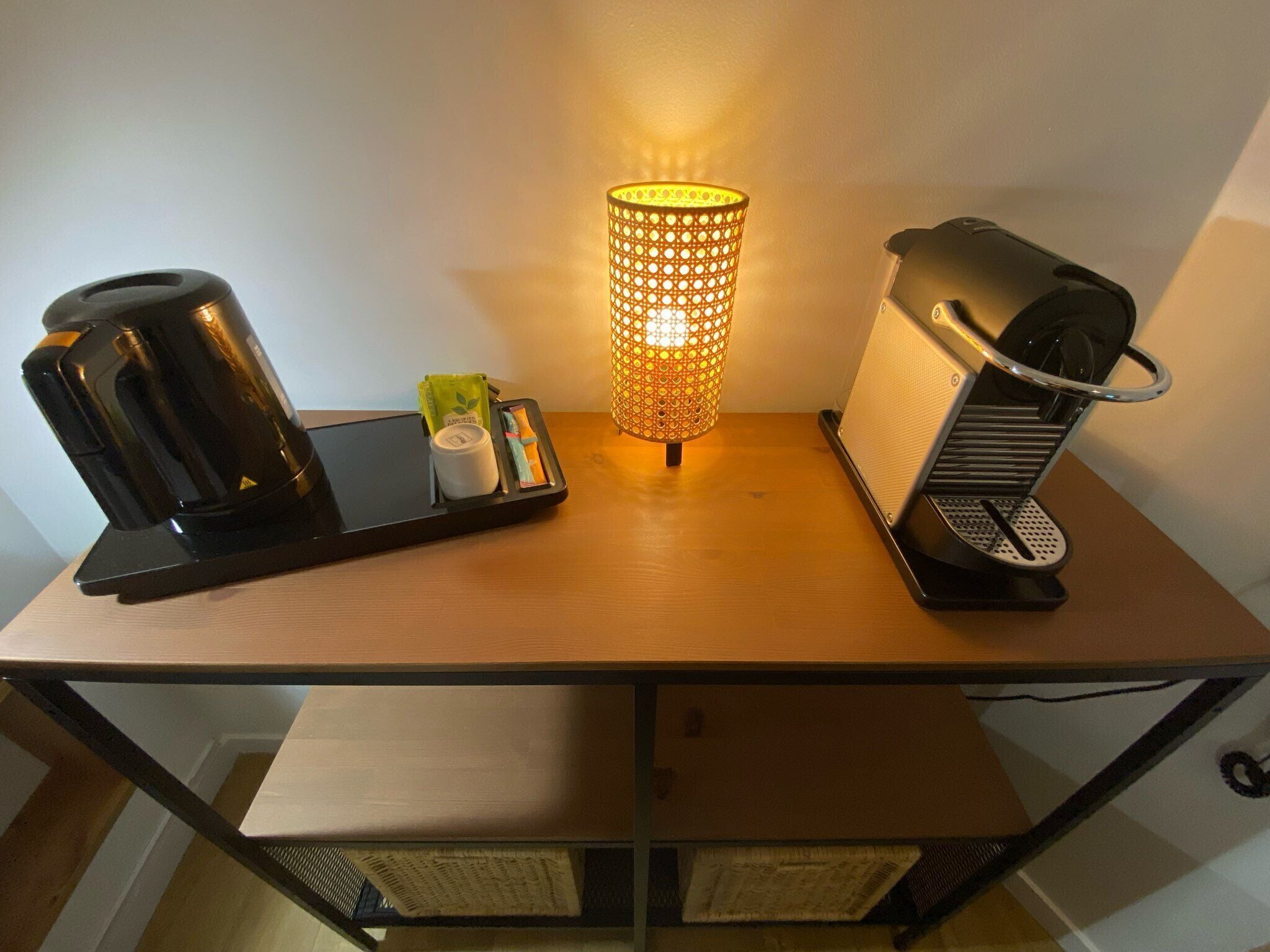 A wooden table with a black electric kettle, a small tray with a cup and a green packet, and a cylindrical lamp with a patterned shade.