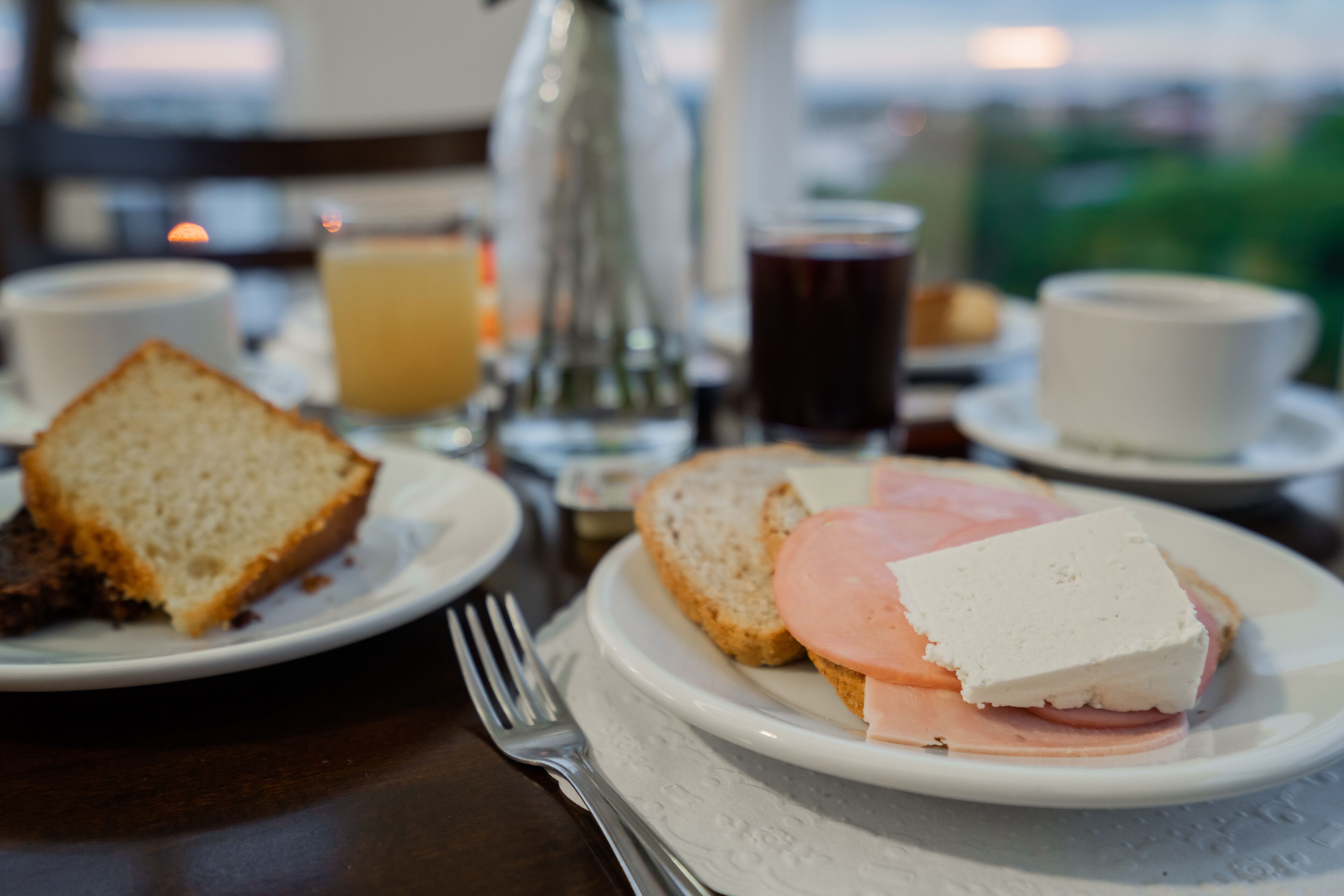Une table mise pour le petit-déjeuner avec des tartines, du jambon et du fromage sur une assiette, une tasse de café et un verre de jus d’orange.