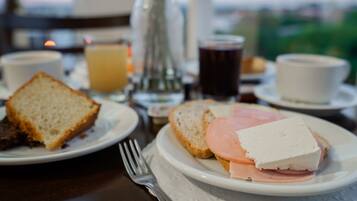 Une table mise pour le petit-déjeuner avec des tartines, du jambon et du fromage sur une assiette, une tasse de café et un verre de jus d’orange.