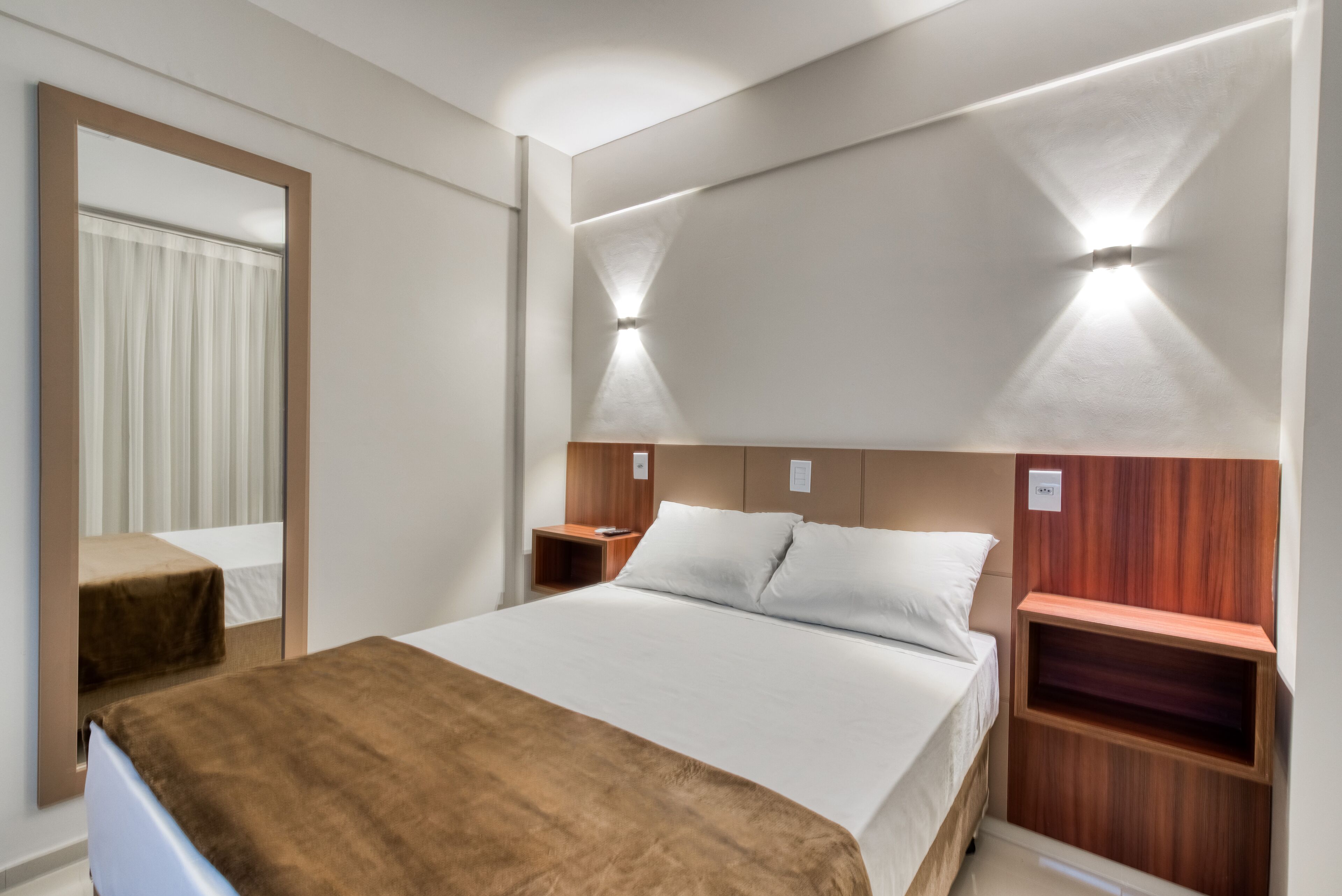 A modern hotel room with a large bed, wooden headboard, bedside tables, and a mirror.