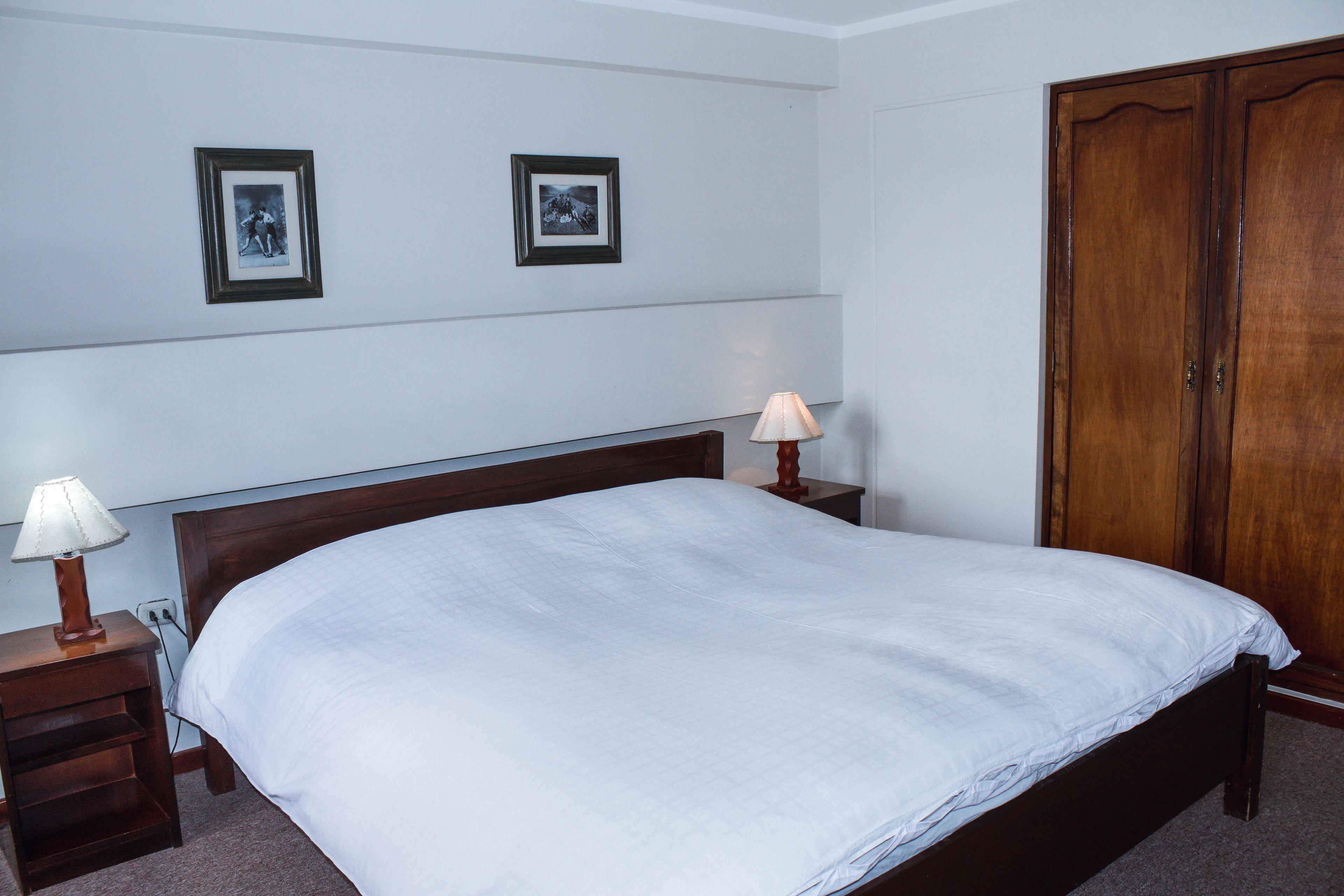 A neatly made bed with a white comforter, a wooden nightstand with a lamp, and a wooden wardrobe.