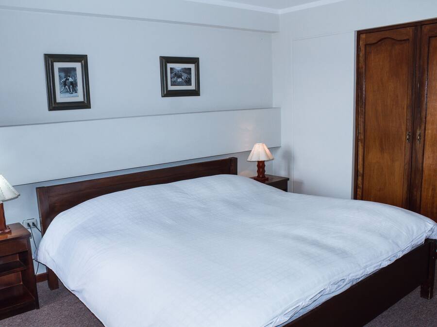 A neatly made bed with a white comforter, a wooden nightstand with a lamp, and a wooden wardrobe.