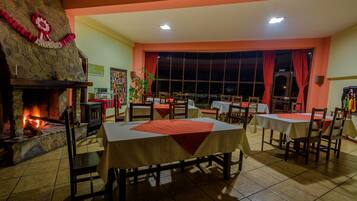 A restaurant with a stone fireplace, multiple tables with red and white tablecloths, and a large window with red curtains.