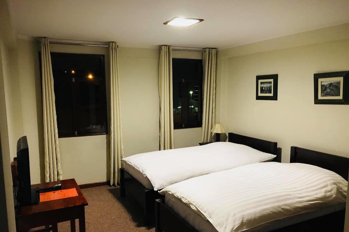A hotel room with a bed, a desk, and a chair. There are two windows with curtains, a lamp on the desk, and framed pictures on the wall.
