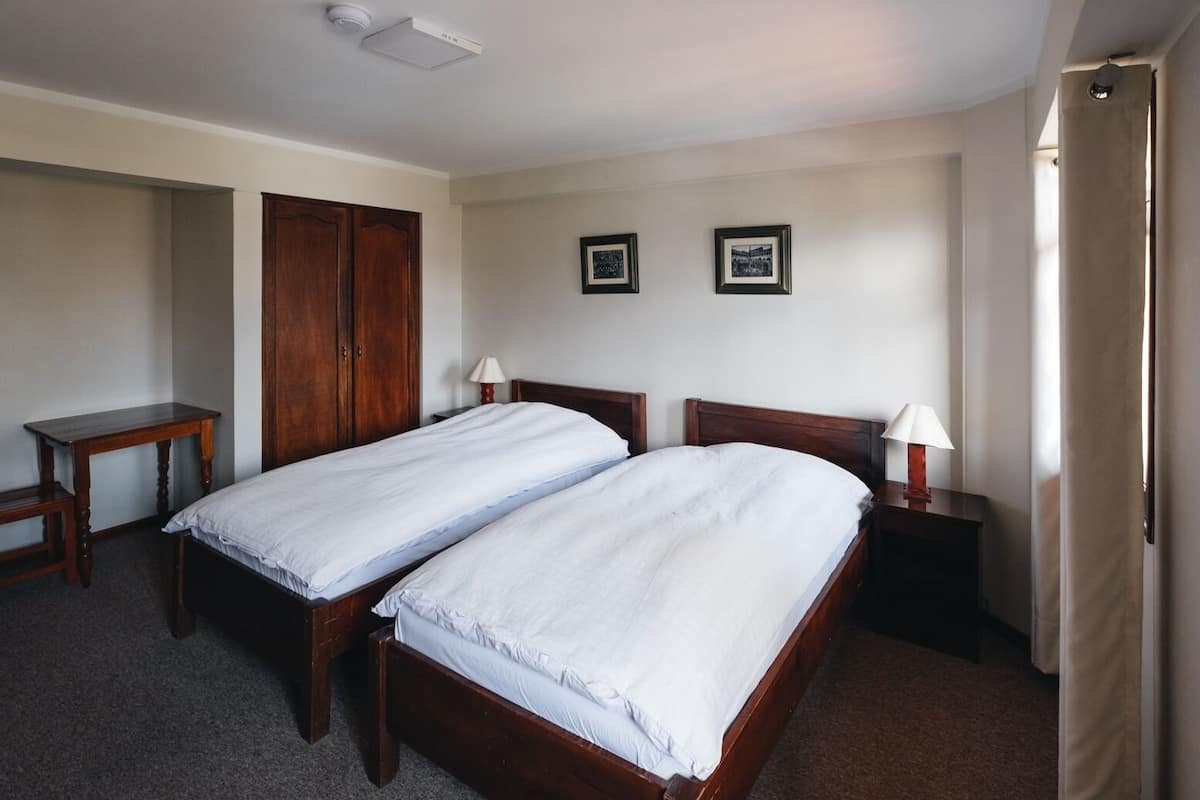 A hotel room with two single beds, a wooden wardrobe, a desk, and two framed pictures on the wall.