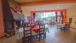 Un comedor con mesas y sillas, una chimenea de piedra con fuego encendido y ventanas grandes con cortinas rojas.