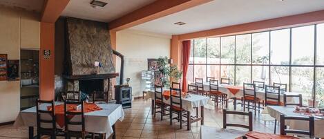 Un restaurante con varias mesas y sillas, una chimenea de piedra y amplios ventanales que ofrecen vistas al exterior.
