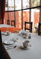 Uma mesa de jantar posta com pratos, talheres, xícara e copo, com uma parede de tijolos e janelas ao fundo.