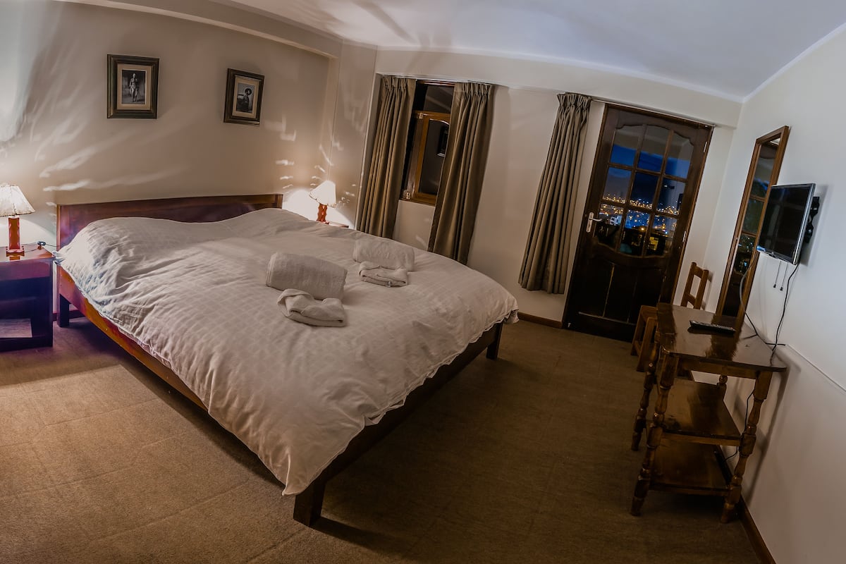 A hotel room with a large bed, two bedside lamps, a desk with a chair, a TV, and a door with curtains.