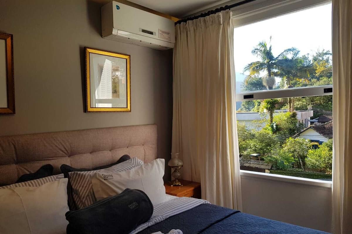 A hotel room with a bed, a framed picture on the wall, an air conditioning unit, and a window with curtains.