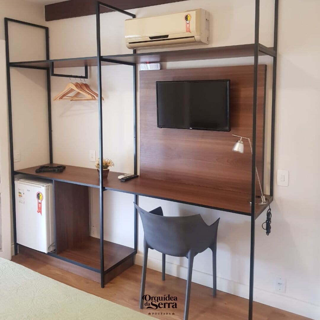 Una habitación con un escritorio de madera, un televisor montado en la pared, una silla, un refrigerador pequeño y un perchero.