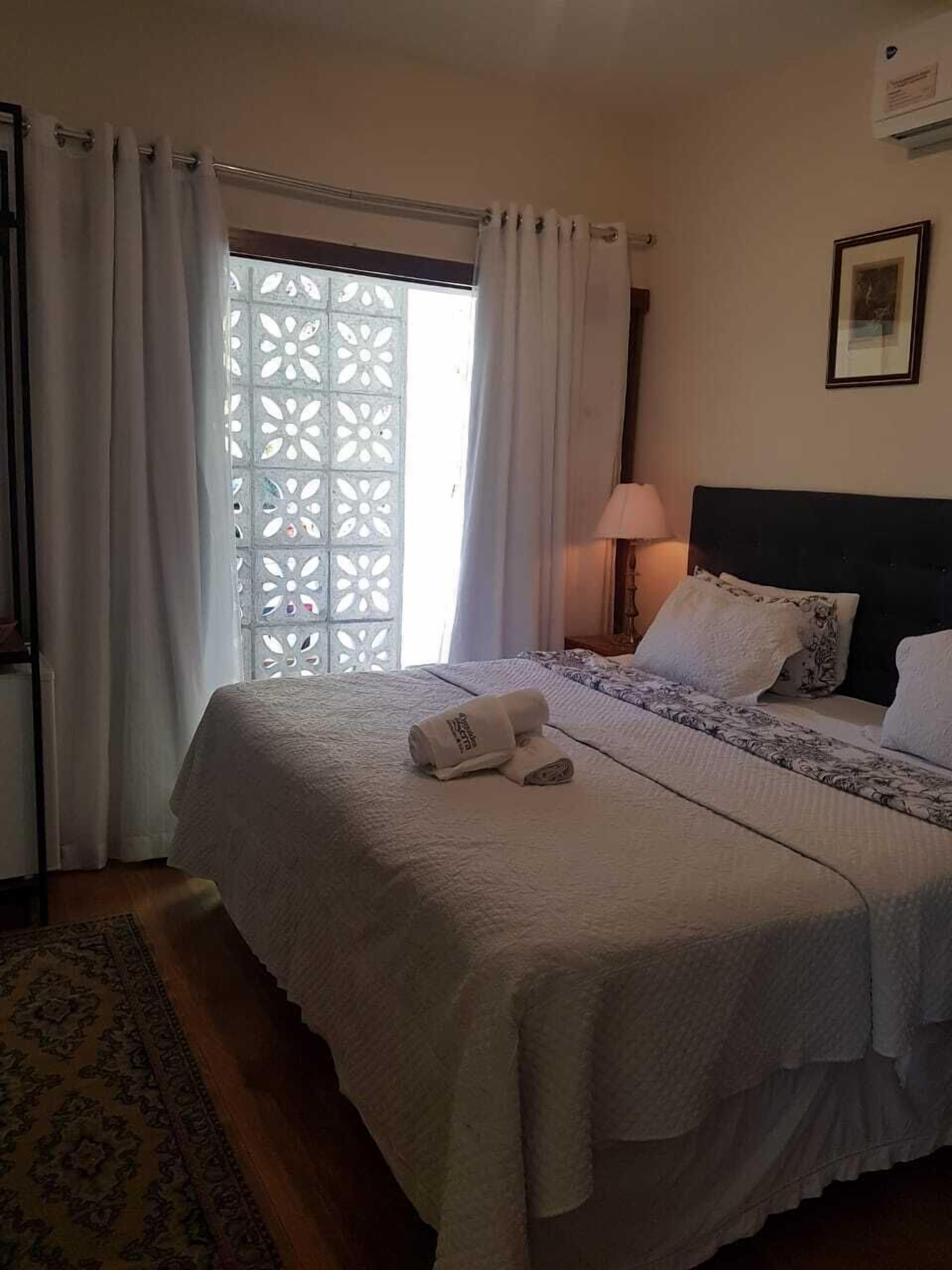 A neatly made bed with white linens, a bedside lamp, and a window with white curtains.