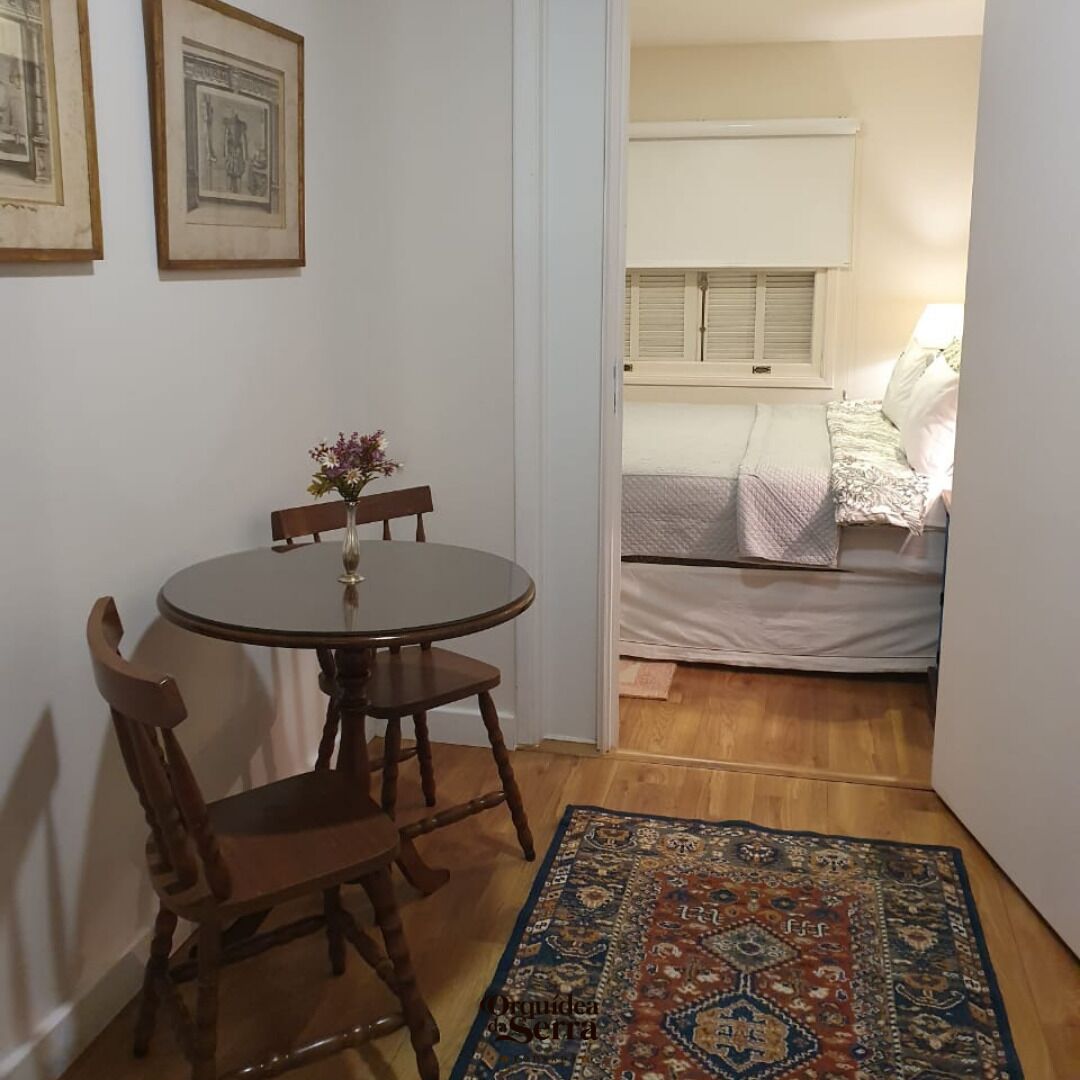 A small room with a round wooden table, two chairs, a vase with flowers, a patterned rug, a bed with a white duvet, and a window with blinds.