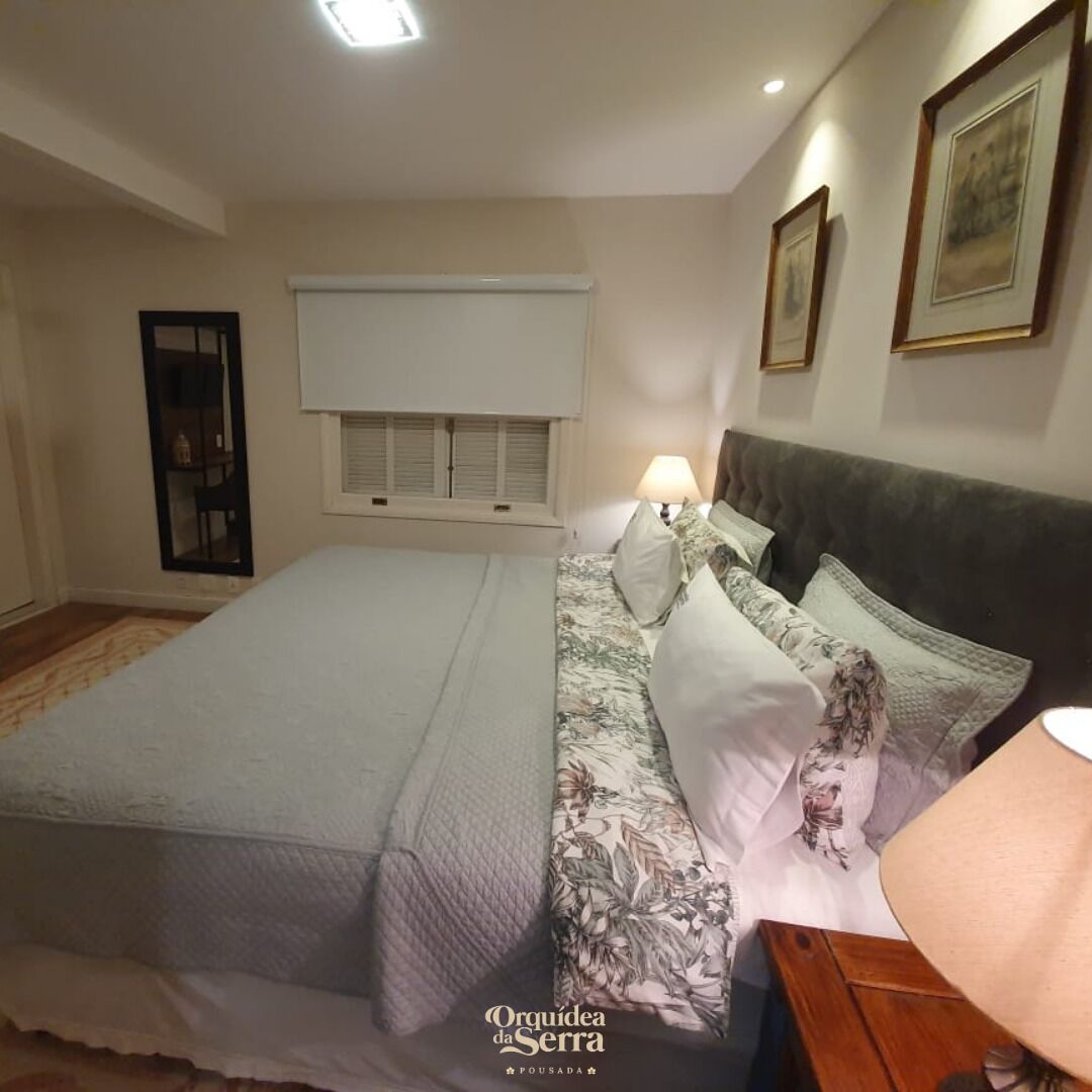 Un dormitorio con cama, mesita de noche, lámpara, ventana con contraventanas y cuadros en la pared.
