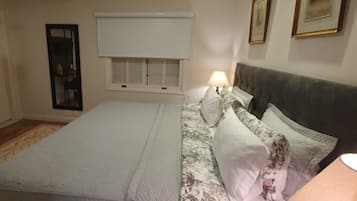 Un dormitorio con cama, mesita de noche, lámpara, ventana con contraventanas y cuadros en la pared.