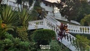A white building with a staircase and a white balustrade, surrounded by lush greenery and trees. - Pousada Orquídea da Serra (Petrópolis)