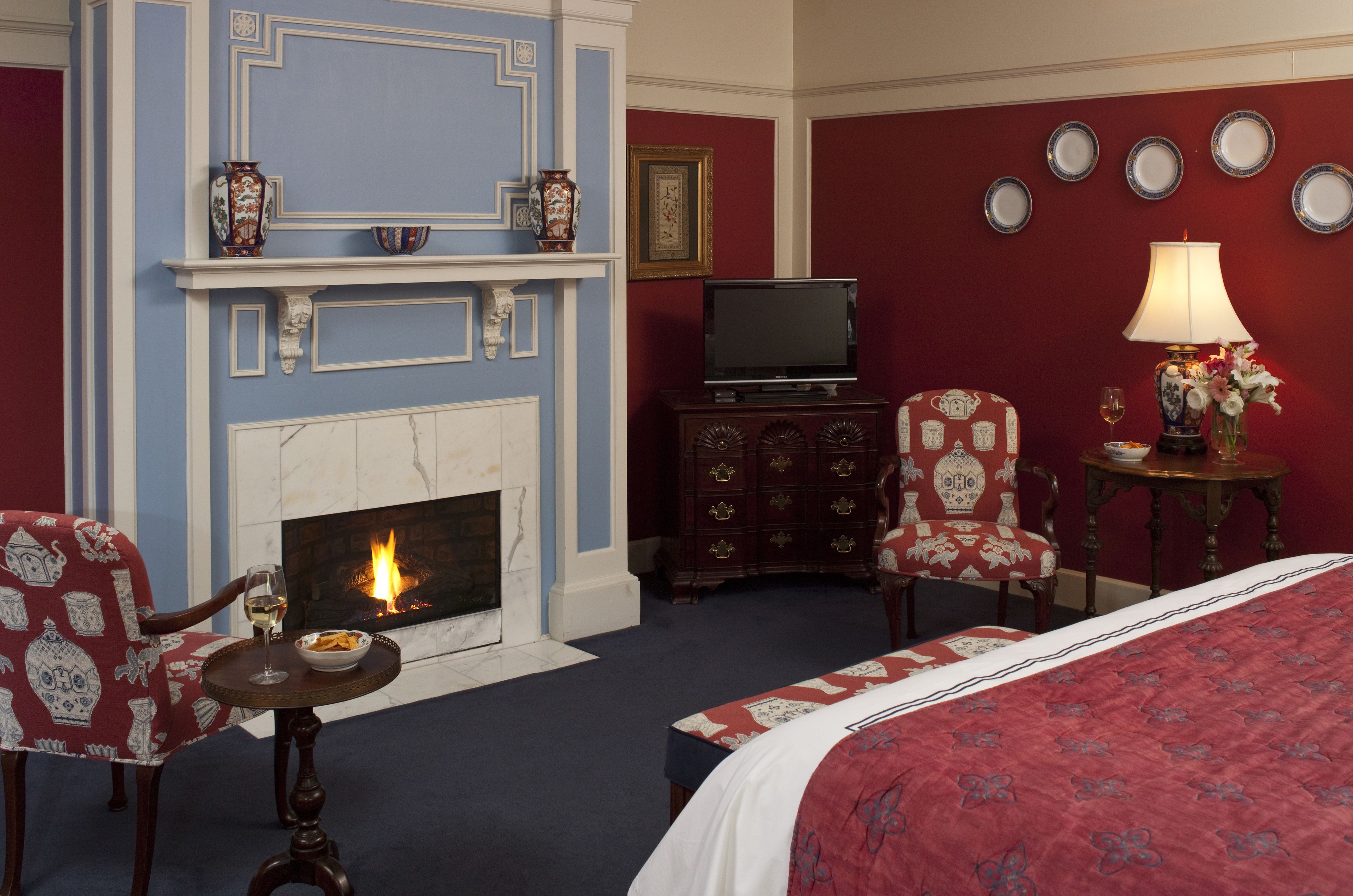 Habitación de hotel con chimenea, televisor, cama, mesa de comedor con una silla y una mesita de noche con lámpara y flores.
