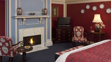 Habitación de hotel con chimenea, televisor, cama, mesa de comedor con una silla y una mesita de noche con lámpara y flores.