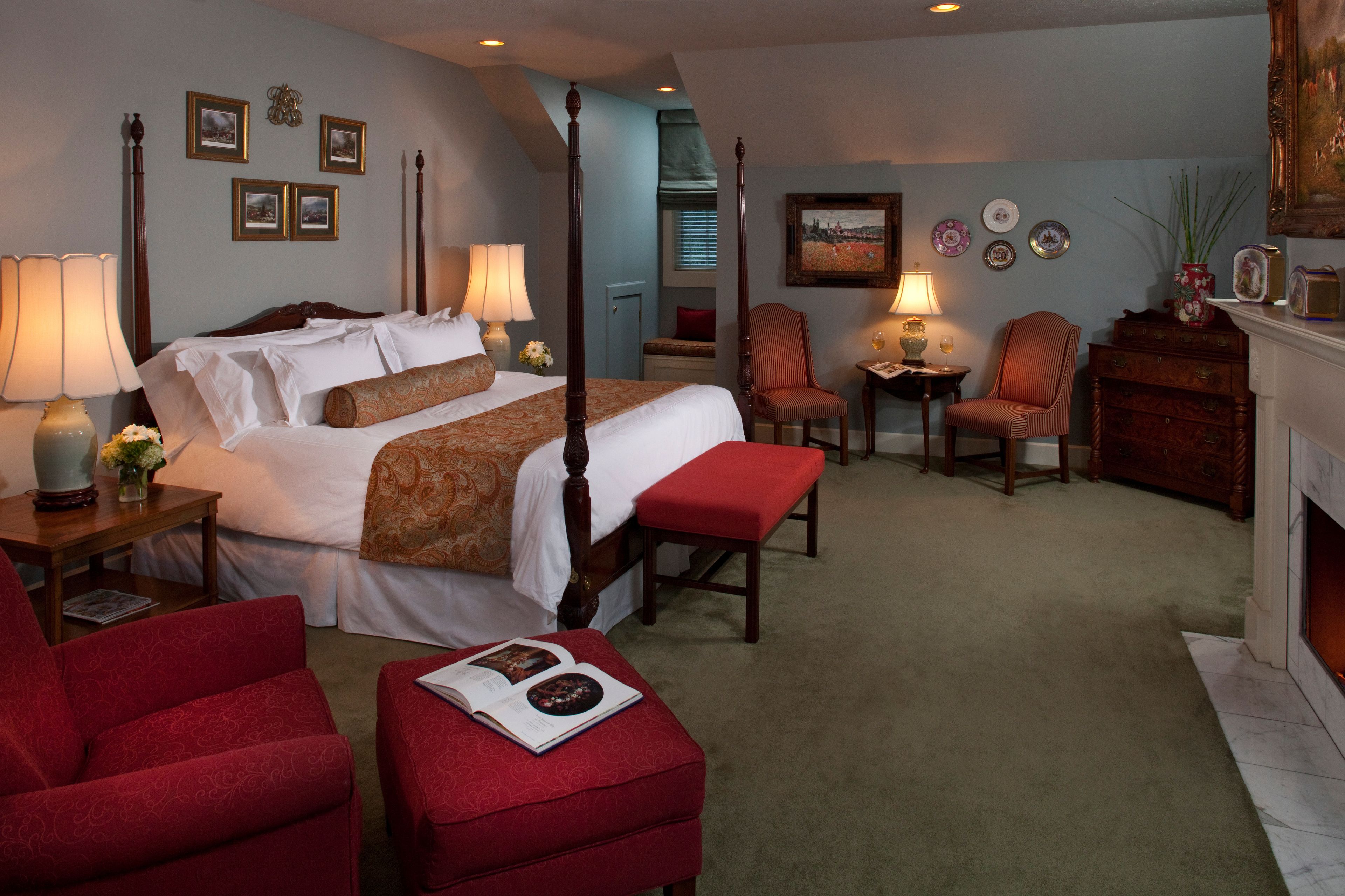 A four-poster bed with a red upholstered bench, a red armchair, and a small table with a lamp.