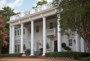 Un edificio grande de estilo colonial, blanco, con un porche cubierto sostenido por columnas, rodeado de setos cuidados y árboles con flores.